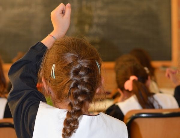 Fotografia alunna scuola primaria di spalle, in classe, con la mano alzata